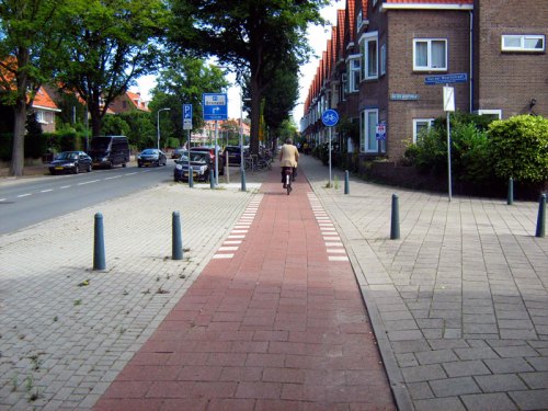 The view of a Dutch-style continuous-path minor junction from the view of a bike rider. The cyclepath and footpath both continue across the junction, and the minor road is disconnected from the main road. Cars have to mount the pavement and cross both paths to get between the two roads.