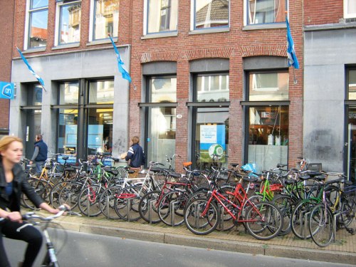 Many bikes parked outside a supermarket in Groningen, Netherlands. A young woman rides past on a bike.